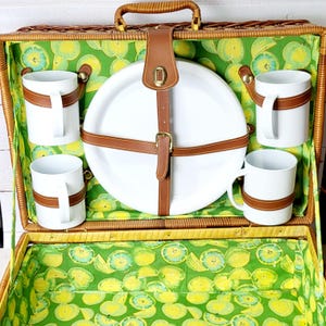 May include: Open picnic basket with a woven exterior, featuring a handle and a green and yellow fruit-patterned interior lining. Includes white mugs with brown leather straps, a white plate secured by brown leather straps, and a matching lid.