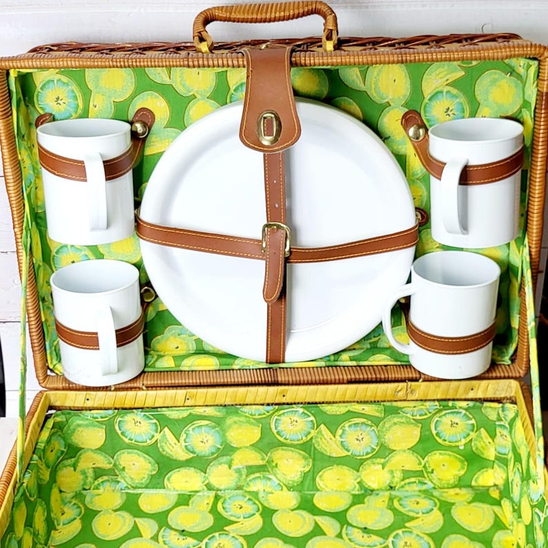 May include: Open picnic basket with a woven exterior, featuring a handle and a green and yellow fruit-patterned interior lining. Includes white mugs with brown leather straps, a white plate secured by brown leather straps, and a matching lid.