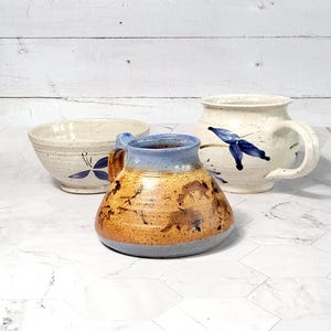 May include: Three handmade ceramic mugs with speckled glaze. One mug is blue and brown with a floral design, another is white with a blue butterfly design, and the third is white with a blue floral design.