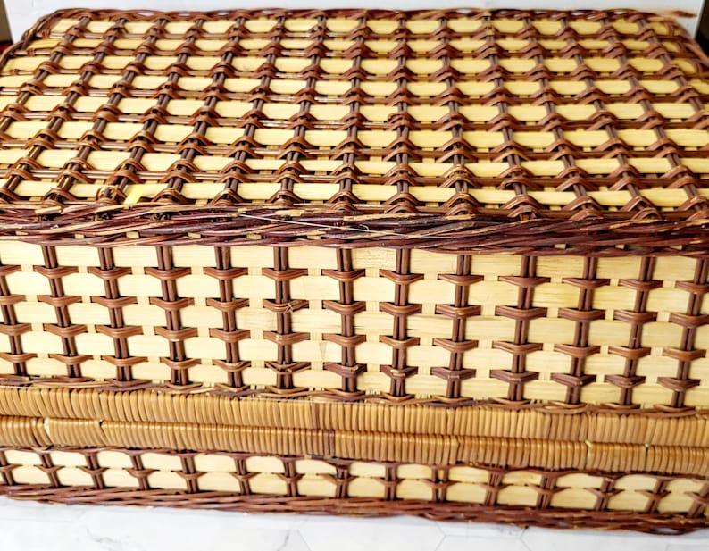 May include: A rectangular picnic basket with a woven design. The basket is made of light brown and dark brown wicker, with a lid and a rectangular shape. The weave pattern is intricate and detailed.