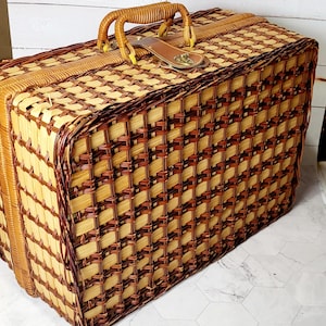 May include: A vintage picnic basket with a woven pattern in shades of light and dark brown. It features a sturdy handle, a leather strap, and a metal clasp. The basket is rectangular and appears to be in good condition.