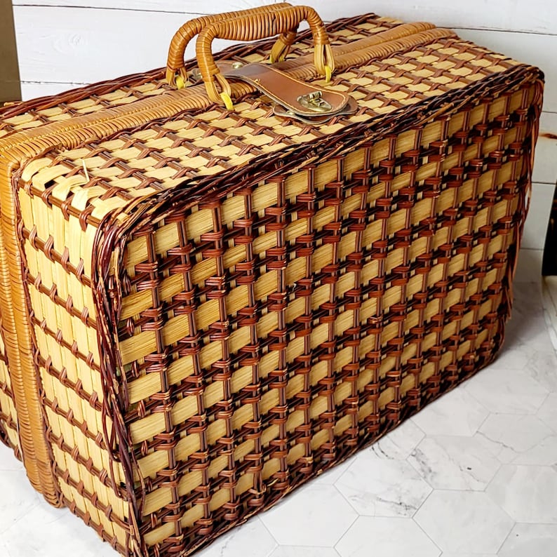 May include: A vintage picnic basket with a woven pattern in shades of light and dark brown. It features a sturdy handle, a leather strap, and a metal clasp. The basket is rectangular and appears to be in good condition.