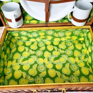 May include: A wicker picnic basket with a green and yellow fruit-patterned interior. The basket contains white mugs with brown leather straps, a white plate, and a matching fruit-patterned lining. The basket is closed with brown leather straps.