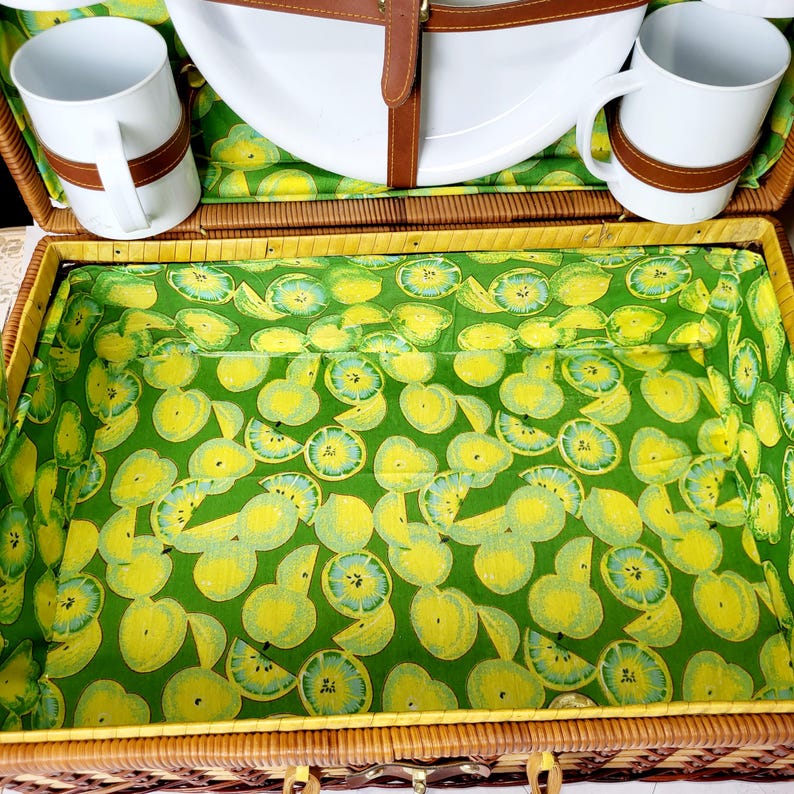 May include: A wicker picnic basket with a green and yellow fruit-patterned interior. The basket contains white mugs with brown leather straps, a white plate, and a matching fruit-patterned lining. The basket is closed with brown leather straps.