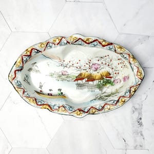 May include: A white ceramic dish with a colourful floral and landscape design. The dish has two sections and a scalloped edge.