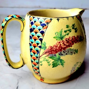 Könnte beinhalten: Eine gelbe Keramikkanne mit einem Blumendesign und einem geometrischen Muster in Blau, Grün, Orange und Weiß. Die Kanne hat einen Henkel und eine craquelée Glasur.