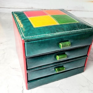 Peut inclure: Une boîte à bijoux en cuir vert avec quatre tiroirs et des accents en cuir coloré sur le dessus. Les tiroirs ont des façades en cuir vert et des boutons en bois.