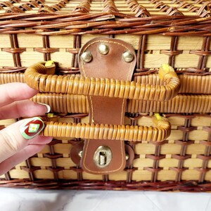 May include: Close-up of a woven wicker picnic basket with a brown leather handle and clasp. The basket is made of light and dark brown wicker, with a woven pattern. The handle is curved and attached to the basket with metal rivets.