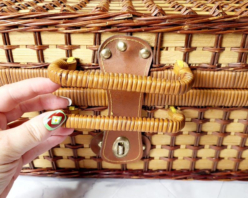 May include: Close-up of a woven wicker picnic basket with a brown leather handle and clasp. The basket is made of light and dark brown wicker, with a woven pattern. The handle is curved and attached to the basket with metal rivets.