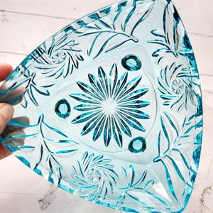Puede incluir: Un plato de vidrio triangular de color azul verdoso con un patrón decorativo de hojas y espirales. El plato tiene tres pequeños pies en la parte inferior.