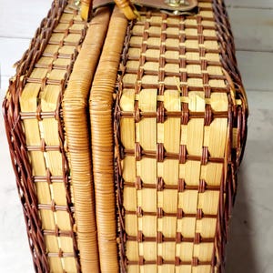 May include: Woven picnic basket with a rectangular shape. The basket features a light brown and dark brown checkered pattern, a brown leather handle, and a metal clasp closure. The sides are reinforced with woven bands.