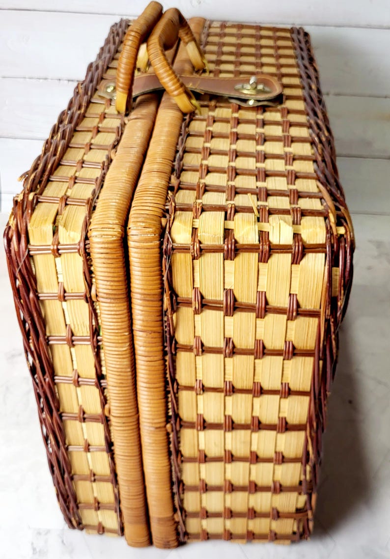 May include: Woven picnic basket with a rectangular shape. The basket features a light brown and dark brown checkered pattern, a brown leather handle, and a metal clasp closure. The sides are reinforced with woven bands.