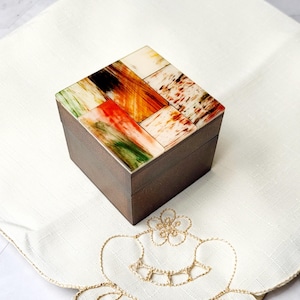 May include: A small wooden box with a colourful, geometric patterned lid. The lid is made of inlaid pieces of wood in shades of brown, orange, green, and white.