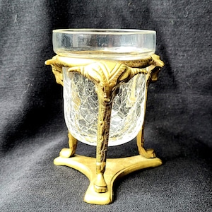 Puede incluir: Un candelero de metal dorado con un portavelas de vidrio transparente. El candelero tiene un diseño decorativo con tres patas y una parte superior redonda. El portavelas de vidrio tiene un acabado craquelado.