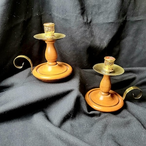 Peut inclure: Deux bougeoirs en bois avec des détails en laiton. Chaque bougeoir a une base ronde, une tige en bois tourné et une coupelle à bougie en laiton. Une poignée incurvée en laiton s'étend sur le côté de chaque base. Les coupelles sont claires et texturées.