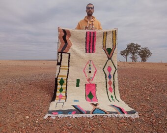 Peut inclure: Un tapis blanc avec des motifs géométriques colorés en rose, bleu, vert et jaune. Le tapis est plié en deux et tenu par une personne.