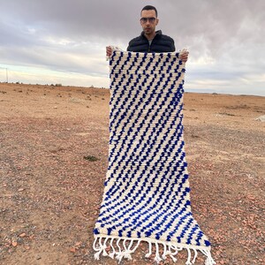 May include: A white and blue striped rug with fringe on the ends. The rug is being held up by a person in a desert landscape.