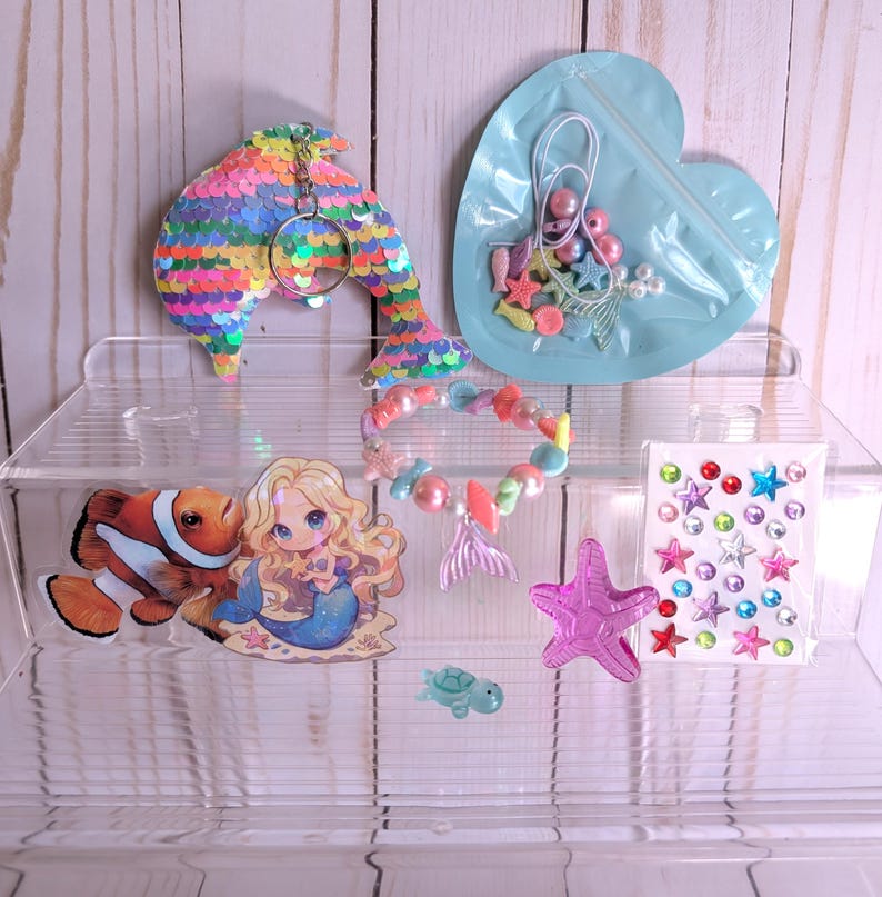 May include: A collection of mermaid-themed craft supplies. Includes a sequined dolphin keychain, a heart-shaped bag with beads and charms, a beaded bracelet, stickers, and a starfish. The items are arranged on a clear acrylic organizer.