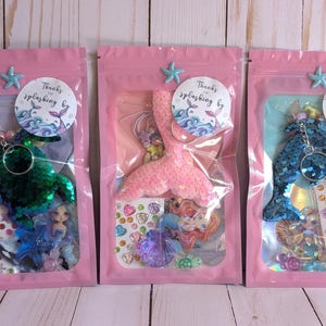 May include: Three pink zipper bags each containing a mermaid-themed keychain, stickers, and small figurines. The keychains are green, pink, and blue, and the bags are decorated with star-shaped embellishments and circular stickers that say "Thanks for splashing by."