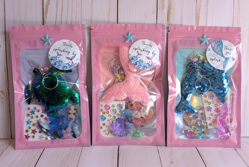 May include: Three pink zipper bags each containing a mermaid-themed keychain, stickers, and small figurines. The keychains are green, pink, and blue, and the bags are decorated with star-shaped embellishments and circular stickers that say "Thanks for splashing by."