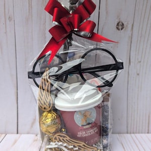 May include: A clear cellophane gift bag topped with a shiny red bow containing black round-rimmed glasses, a small cup of color-changing hot chocolate mix, a gold-wrapped Lindor chocolate truffle, and two wooden wing-shaped cutouts.