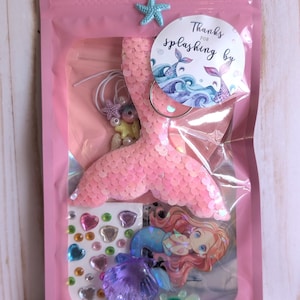 May include: A pink cellophane bag containing mermaid-themed craft supplies. Includes a sequined pink mermaid tail, stickers, a mermaid charm, and a sticker that says "Thanks for splashing by". A blue starfish decorates the top.