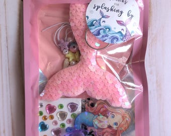 May include: A pink cellophane bag containing mermaid-themed craft supplies. Includes a sequined pink mermaid tail, stickers, a mermaid charm, and a sticker that says "Thanks for splashing by". A blue starfish decorates the top.