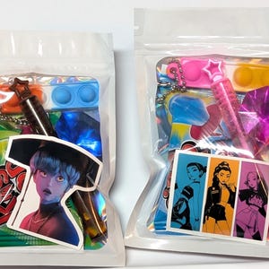 May include: Two clear plastic bags filled with colorful items. One bag contains a sticker of a person with blue skin and a black hat, a blue pop-it, and a star-shaped keychain. The other bag has a sticker with three women, a pink pop-it, and a pink star-shaped pen.
