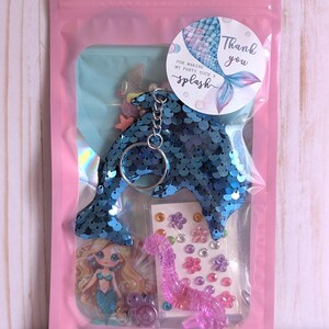 May include: A pink cellophane bag contains a mermaid-themed party favor set. The set includes a blue sequined dolphin keychain, stickers, and a mermaid-themed card. A round sticker with a mermaid tail and the words "Thank you" is also included.