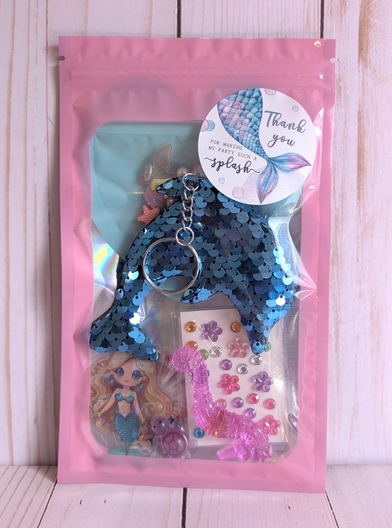 May include: A pink cellophane bag contains a mermaid-themed party favor set. The set includes a blue sequined dolphin keychain, stickers, and a mermaid-themed card. A round sticker with a mermaid tail and the words "Thank you" is also included.