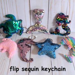 May include: A collection of sequined keychains in various shapes, including a turtle, mermaid tail, mermaid, dolphin, seahorse, and starfish. The keychains are in a variety of colors, including pink, blue, green, and rainbow. The text "flip sequin keychain" is at the bottom.