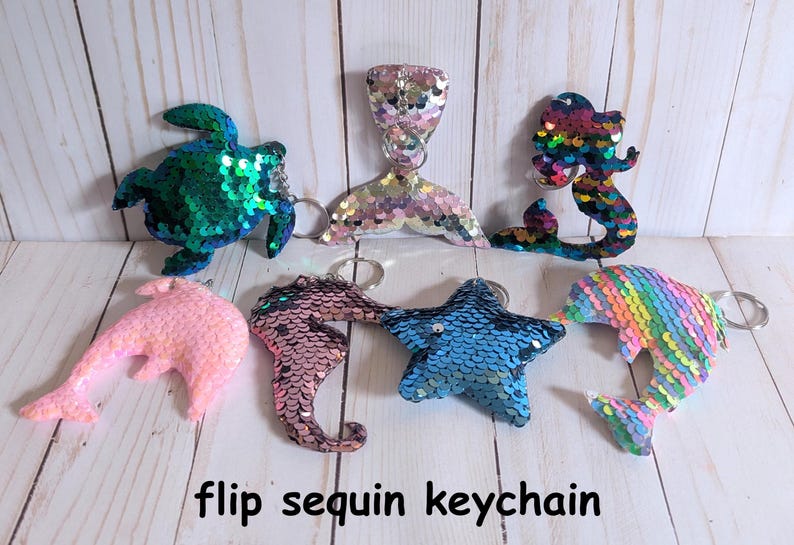 May include: A collection of sequined keychains in various shapes, including a turtle, mermaid tail, mermaid, dolphin, seahorse, and starfish. The keychains are in a variety of colors, including pink, blue, green, and rainbow. The text "flip sequin keychain" is at the bottom.