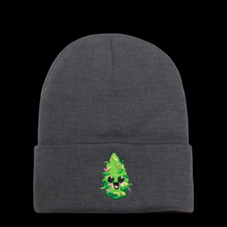 May include: A dark gray beanie with a green cartoon marijuana leaf embroidered on the front. The leaf has a smiling face with black eyes and a pink mouth.