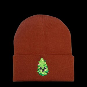 May include: A brown beanie with a green marijuana leaf design. The leaf has a smiling face with black eyes.