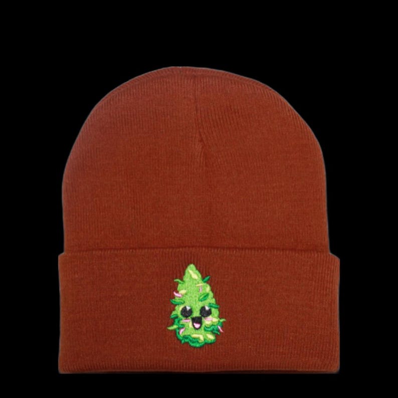 May include: A brown beanie with a green marijuana leaf design. The leaf has a smiling face with black eyes.