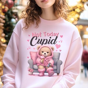 May include: Pink sweatshirt with the text "Not Today Cupid." The graphic features a teddy bear wearing a pink hooded outfit, holding an ice cream cone and a remote control, seated on a gray couch. Pink hearts and musical notes are also included.