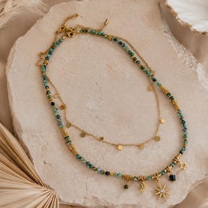 May include: A layered necklace set featuring one strand of small turquoise-colored beads interspersed with gold-toned cylindrical spacers and a second delicate gold-toned chain with small circular discs, a crescent moon, and a star charm.