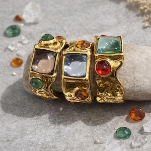 May include: Three gold-toned rings with large, colorful gemstones. The rings feature square and round stones in shades of pink, blue, green, and red. The rings are displayed on a gray stone surface.