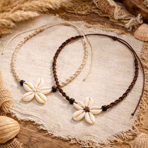 May include: Two seashell necklaces. One is tan, the other is brown. Each necklace features a flower made of white seashells and dark beads. The necklaces are displayed on a beige cloth, with seashells and dried plants in the background.