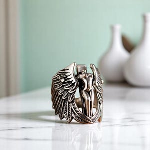 Könnte beinhalten: Ein silberner Ring mit einem detaillierten Design einer geflügelten Frau. Der Ring liegt auf einer weißen Marmorfläche.