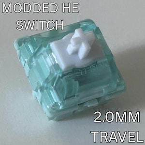 Pode incluir: Uma imagem em close-up de um interruptor HE modificado com uma caixa transparente azul clara e uma haste branca em forma de cruz. O texto "MODDED HE SWITCH" e "2.0MM TRAVEL" são exibidos em branco.