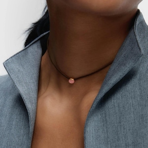 May include: A necklace with a small, round, pink gemstone pendant on a thin, dark brown cord. The person is wearing a blue denim jacket. The pendant is a focal point.