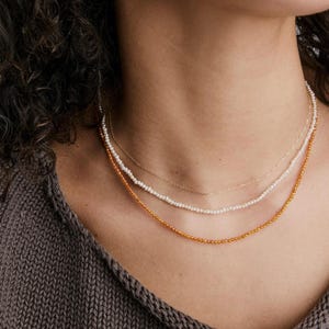 May include: Three layered necklaces. The top is a fine gold chain. The middle necklace has small white pearls. The bottom necklace is made of small, amber-coloured beads. The necklaces are worn around the neck.