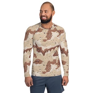 Puede incluir: Camiseta de manga larga con un estampado de camuflaje desértico en tonos beige, marrón y blanco. La camiseta tiene cuello redondo y mangas raglán. El modelo lleva pantalones cortos azules.