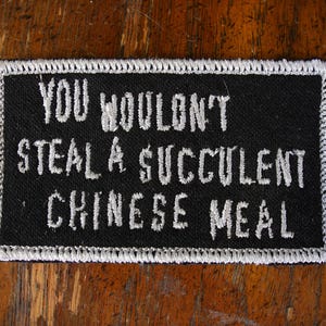 May include: Black rectangular patch with white embroidered text: "YOU WOULDN'T STEAL A SUCCULENT CHINESE MEAL." The patch has a white border and is set against a wooden background. A humorous statement.