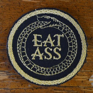 Może przedstawiać: Naszywka haftowana w kolorze czarnym i złotym z wzorem węża i tekstem "EAT ASS".