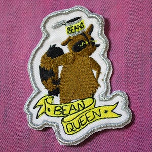 Könnte beinhalten: Gestickter Aufnäher mit einem Cartoon-Waschbären, der einen "Beans"-Hut trägt. Der Waschbär ist braun mit schwarzer Maske und gestreiftem Schwanz. Ein gelbes Banner darunter zeigt "Bean Queen". Weißer Rand mit silbernen Nähten.