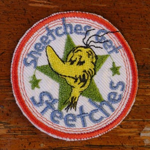 May include: A white, round, embroidered patch with a red and orange border. The patch features a green star with a yellow creature with a long nose and a black eye. The text "Stretches Stretches" is written in blue around the star.