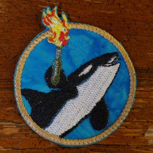 Könnte beinhalten: Eine gestickte Orca in Schwarzweiß mit einer gelben und roten Flamme oben drauf, alles in einem goldenen Kreis auf blauem Hintergrund.