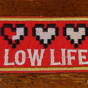 Peut inclure: Patch brodé rouge avec trois cœurs pixélisés et le texte "LOW LIFE" en blanc.